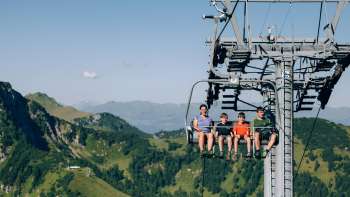 Una famiglia sale sulla seggiovia Sareis in un paesaggio montano verdeggiante