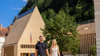 Una coppia passeggia nella soleggiata Piazza del Governo di Vaduz, con la vista del moderno edificio del Parlamento e del Castello di Vaduz sullo sfondo.