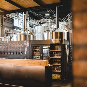 Vista interna di una birreria con impianto di produzione in acciaio inox e invitante bar in primo piano.