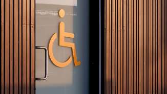 Simbolo arancione della sedia a rotelle su una porta di vetro che indica una toilette accessibile, con pannelli di legno sullo sfondo.