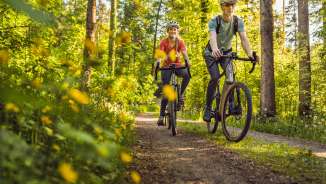 Due ciclisti con casco pedalano rilassati sulle loro bici da ghiaia su un sentiero forestale soleggiato attraverso una tettoia verde - vacanze attive nella natura del Liechtenstein