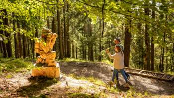 Due escursionisti passano davanti a una figura di legno sul «sentiero delle leggende Walser»