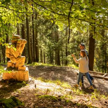 Una donna indica una dettagliata scultura in legno nella foresta del «sentiero delle leggende Walser»