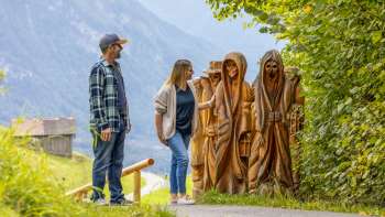 Due escursionisti guardano le figure di legno intagliate sul «sentiero delle leggende Walser» un percorso tematico nel mezzo di un paesaggio alpino con vista sulle montagne Due escursionisti guardano le figure di legno intagliate sul «sentiero delle leggende Walser» un percorso tematico nel mezzo di un paesaggio alpino con vista sulle montagne