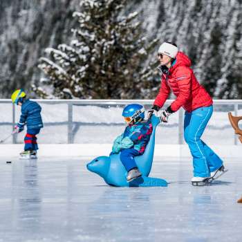Bambini sulla pista di ghiaccio punto di ritrovo Schlucher-Treff a Malbun, sostenuti da un ausilio per il pattinaggio a forma di animale blu.