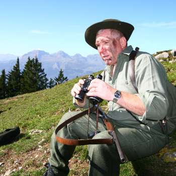 Un cacciatore con un binocolo siede in un prato sullo sfondo di una montagna alpina.