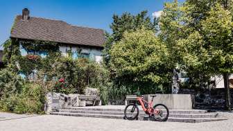 Una e-bike rossa davanti a un edificio storico del Liechtenstein, circondato da alberi e fontane, punto di partenza ideale per un tour in bicicletta attraverso il paese