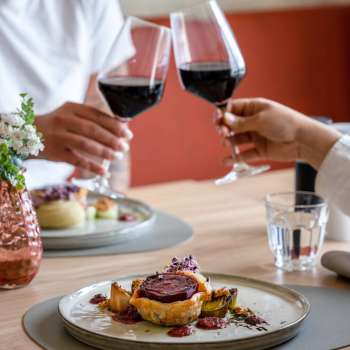 Due persone brindano con vino rosso, in primo piano un piatto principale preparato con cura