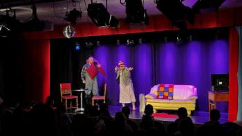 Scena di cabaret al K-BUM in Liechtenstein - palco colorato con divano, duo di attori e pubblico in sala.