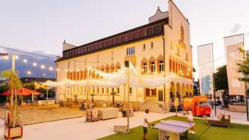 La piazza del municipio di Vaduz di sera, durante un'intensa giornata di mercato, con folla di persone, bancarelle e luci d'atmosfera.