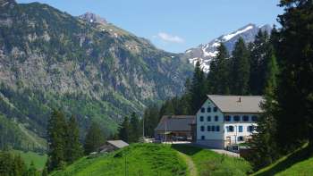 Tradizionale locanda alpina e di montagna Sücka nel Liechtenstein, immersa in un verde paesaggio montano con vista su pendii boscosi e cime innevate. Tradizionale locanda alpina e di montagna Sücka nel Liechtenstein, immersa in un verde paesaggio montano con vista su pendii boscosi e cime innevate.