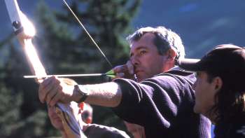 Un uomo si concentra sull'arco durante il tiro con l'arco a Malbun, mentre una guida lo affianca: un'emozionante esperienza all'aperto in mezzo alle montagne.