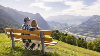 Due escursionisti siedono su una panchina e si godono la vista sulla valle del Reno