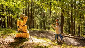 Due escursionisti passano davanti a una figura di legno sul «sentiero delle leggende Walser»