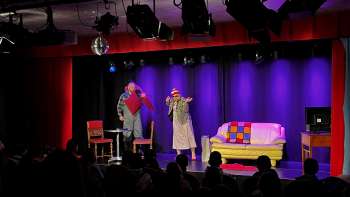 Scena di cabaret al K-BUM in Liechtenstein - palco colorato con divano, duo di attori e pubblico in sala.