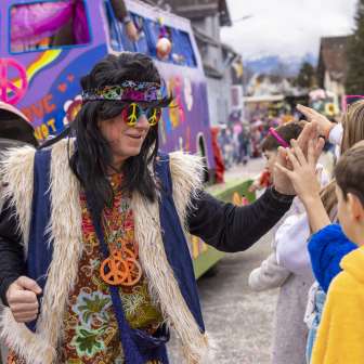 Hippie travestito al carnevale dei bambini di Mauren, dà il cinque ai bambini davanti a un carro colorato.