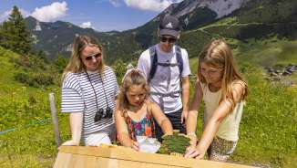 La famiglia esplora una stazione interattiva sul sentiero tematico di Malbun, circondato da paesaggi alpini e prati estivi nel Liechtenstein.