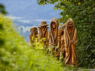 Vista dettagliata delle figure di legno intagliate in una rappresentazione mistica sul Sentiero della Leggenda Walser a Triesenberg, circondato da una natura verdeggiante