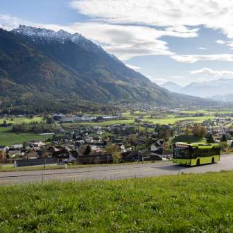 Autobus LIEmobil verde che percorre una strada tortuosa sopra il Liechtenstein con vista sulla valle del Reno e sulle Alpi circostanti