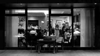 Foto in bianco e nero di un ristorante affollato con vista dall'esterno attraverso la facciata in vetro.