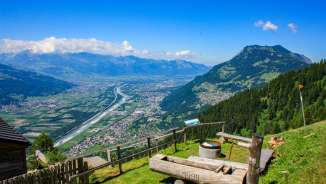 Piattaforma panoramica con fontana in legno e vista sulla valle del Reno e sui comuni del Liechtenstein in una limpida giornata estiva.