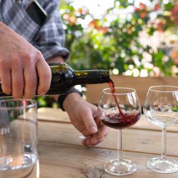 Un bicchiere di vino rosso viene versato da una bottiglia in un bicchiere da vino, con piante fiorite sullo sfondo.
