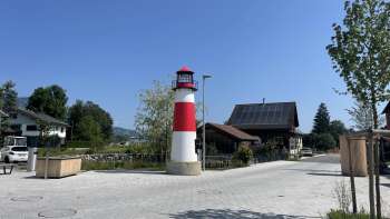 Piccolo faro Moby rosso e bianco su una piazza di Ruggell con tempo soleggiato, con edifici tradizionali sullo sfondo.