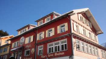Vista esterna dell'Alpenhotel Vögeli Malbun con l'Elchbar al piano terra - edificio tradizionale rosso con scritte bianche contro un cielo blu.