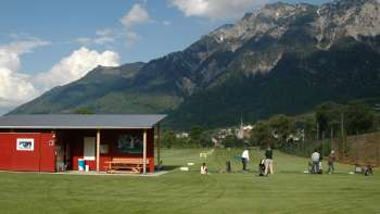 Giocatori di golf sul campo pratica in Liechtenstein con vista su prati verdi, una clubhouse rossa e il paesaggio montano circostante sotto un cielo azzurro.
