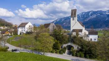 Vista panoramica della «Kirchhügel» (collina della chiesa) di Bendern con la sua chiesa storica, circondata da sentieri curati, alberi e architettura tradizionale sullo sfondo delle montagne alpine.