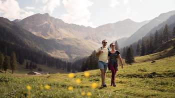 Due persone che attraversano un ampio prato alpino con vista sulle montagne circostanti dell'alpe Valüna con tempo soleggiato