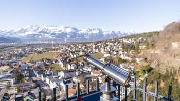 Telescopio presso il punto panoramico Schlossweg Känzile con vista su Vaduz e il panorama alpino innevato sullo sfondo.