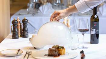 Piatto gourmet servito con carne e contorno, disposto in modo decorativo sotto una cloche bianca nell'elegante ristorante Alpspitz.