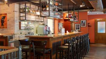 bar del Nuovo Castello di Vaduz con un lungo bancone in legno, sgabelli alti e scaffali in vetro appesi in un ambiente caldo.