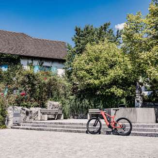 Una e-bike rossa davanti a un edificio storico del Liechtenstein, circondato da alberi e fontane, punto di partenza ideale per un tour in bicicletta attraverso il paese