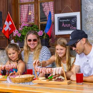 Famiglia seduta su un soleggiato pascolo alpino nel Liechtenstein a gustare una merenda tradizionale con specialità regionali e un panorama montano