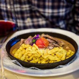 Spaetzle abbondanti con ragù di manzo, guarniti con pomodoro, serviti in padella - accompagnati da un bicchiere di vino rosso su una tavola imbandita.