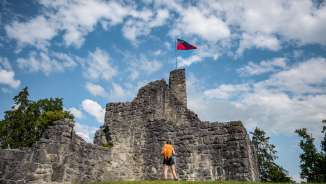 Escursionista davanti alle rovine storiche del castello di Schellenberg con la bandiera del Liechtenstein che sventola sotto un cielo azzurro