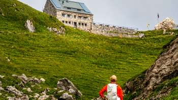 Escursionista con zaino rosso che guarda verso il rifugio Pfälzerhütte nel mezzo di un verde paesaggio montano.