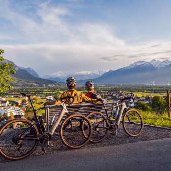Due ciclisti con il casco si siedono su una panchina sopra la valle del Reno e si godono il tramonto con vista sulle Alpi - un'attrazione del tour ciclistico in Liechtenstein
