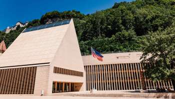 Moderno edificio del parlamento di Vaduz con timpano a punta, bandiera del Liechtenstein di fronte e circondato da verdi colline.