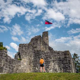 Escursionista davanti alle rovine storiche del castello di Schellenberg con la bandiera del Liechtenstein che sventola sotto un cielo azzurro