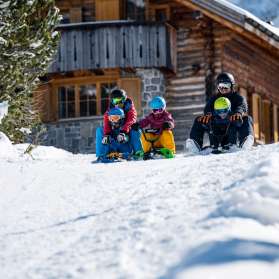 Due adulti e tre bambini siedono sulle slitte e iniziano una discesa insieme davanti a una baita innevata a Malbun
