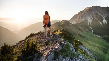 Escursionista con zaino in piedi sul crinale di una montagna del Liechtenstein e sguardo rivolto al sole nascente.