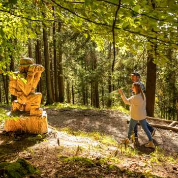 Una donna indica una dettagliata scultura in legno nella foresta del «sentiero delle leggende Walser»