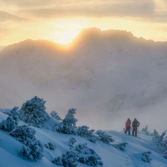 Due persone, tra cui Tina Weirather, camminano attraverso un paesaggio invernale innevato al tramonto sulle montagne del Liechtenstein.