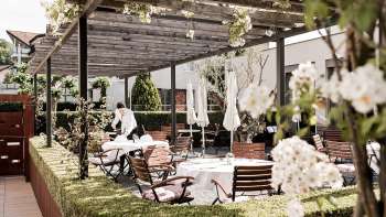 Un'elegante terrazza con tavoli bianchi apparecchiati con tovaglioli e bicchieri sotto un padiglione di legno, circondato da una vegetazione lussureggiante.