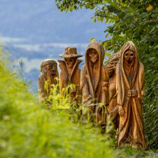 Vista dettagliata delle figure di legno intagliate in una rappresentazione mistica sul Sentiero della Leggenda Walser a Triesenberg, circondato da una natura verdeggiante