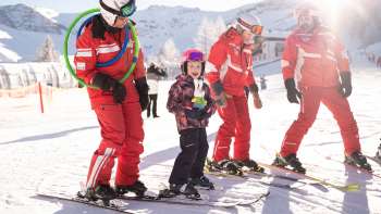 Tre maestri di sci in tuta rossa e un bambino sugli sci nella neve, montagne innevate e sole sullo sfondo