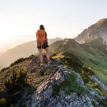 Escursionista con zaino in piedi sul crinale di una montagna del Liechtenstein e sguardo rivolto al sole nascente.